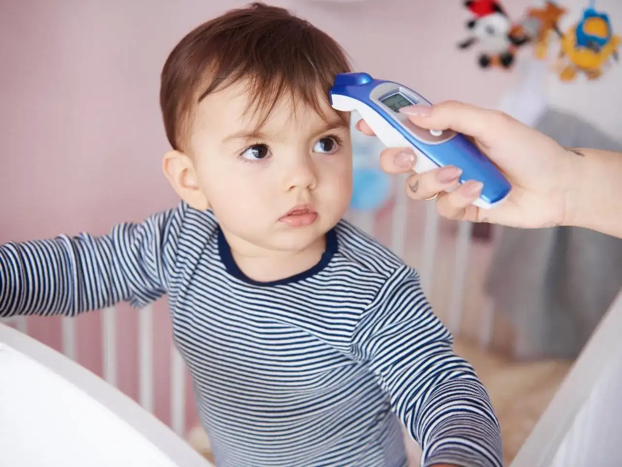 Mama mierzy temperaturę niemowlaka termometrem bezdotykowym. Dziecko ma na sobie pasiasty pajacyk.