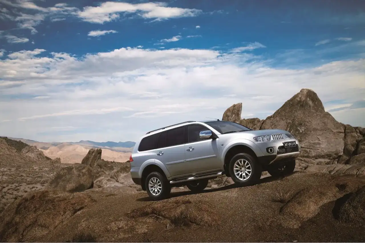 Srebrny Mitsubishi Pajero Sport pokonuje skalisty teren na tle pustynnego krajobrazu i błękitnego nieba z chmurami.