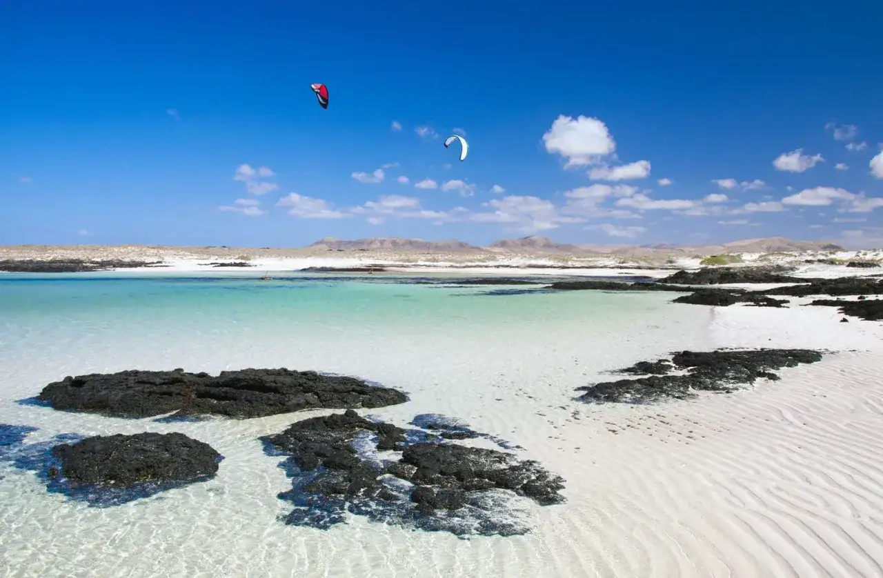 Aguas turquesas y arena blanca en Fuerteventura, con kitesurfistas en el horizonte. Un paraíso entre los 10 sitios para visitar en Fuerteventura.