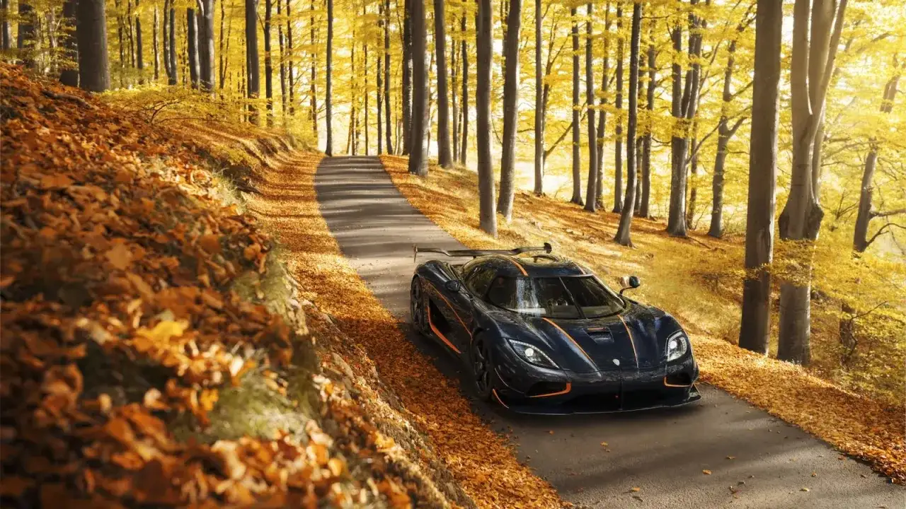 Mroczny Koenigsegg Agera RS z pomarańczowymi akcentami przemierza jesienną drogę wśród złotych liści i drzew.