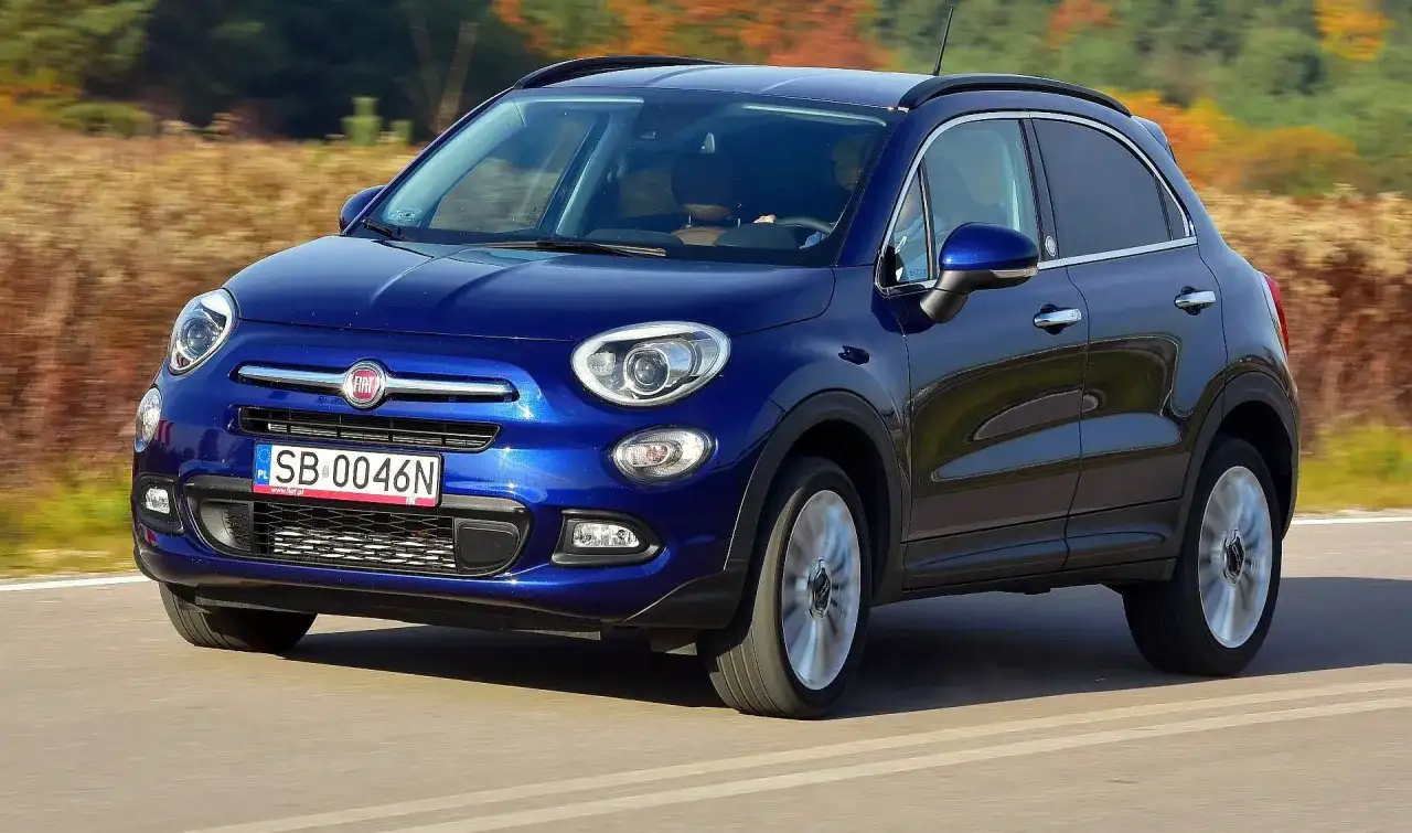 Błękitny Fiat 500X jedzie po drodze, otoczony jesienną przyrodą.