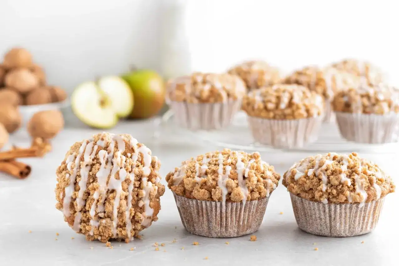 Leckere Apfelmuffins mit Streuseln und Zuckerguss, perfekt für ein Apfelmuffins Rezept. Daneben liegen Äpfel und Zimtstangen.