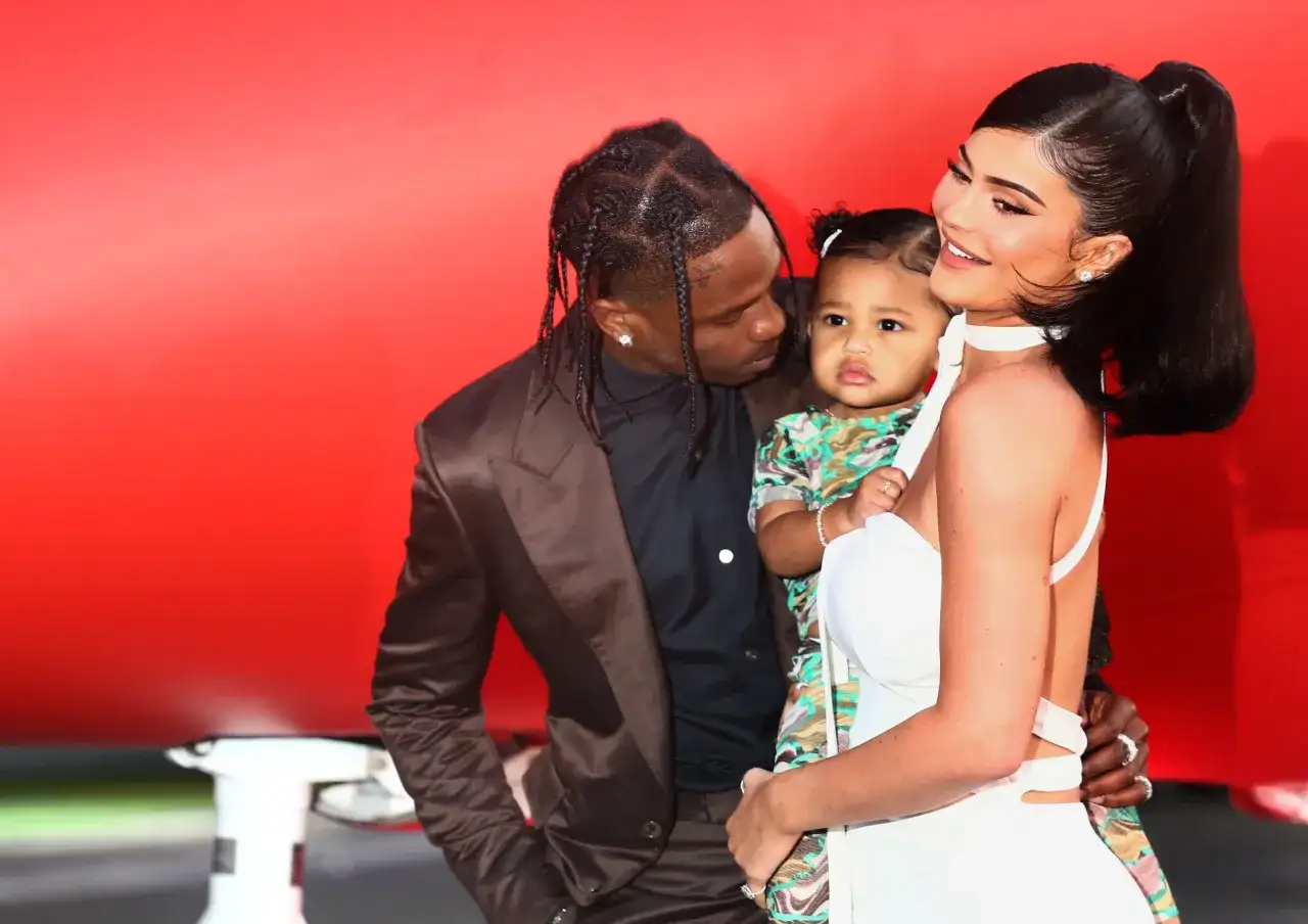 Kylie Jenner z dzieckiem i Travisem Scottem na tle czerwonej ściany.
