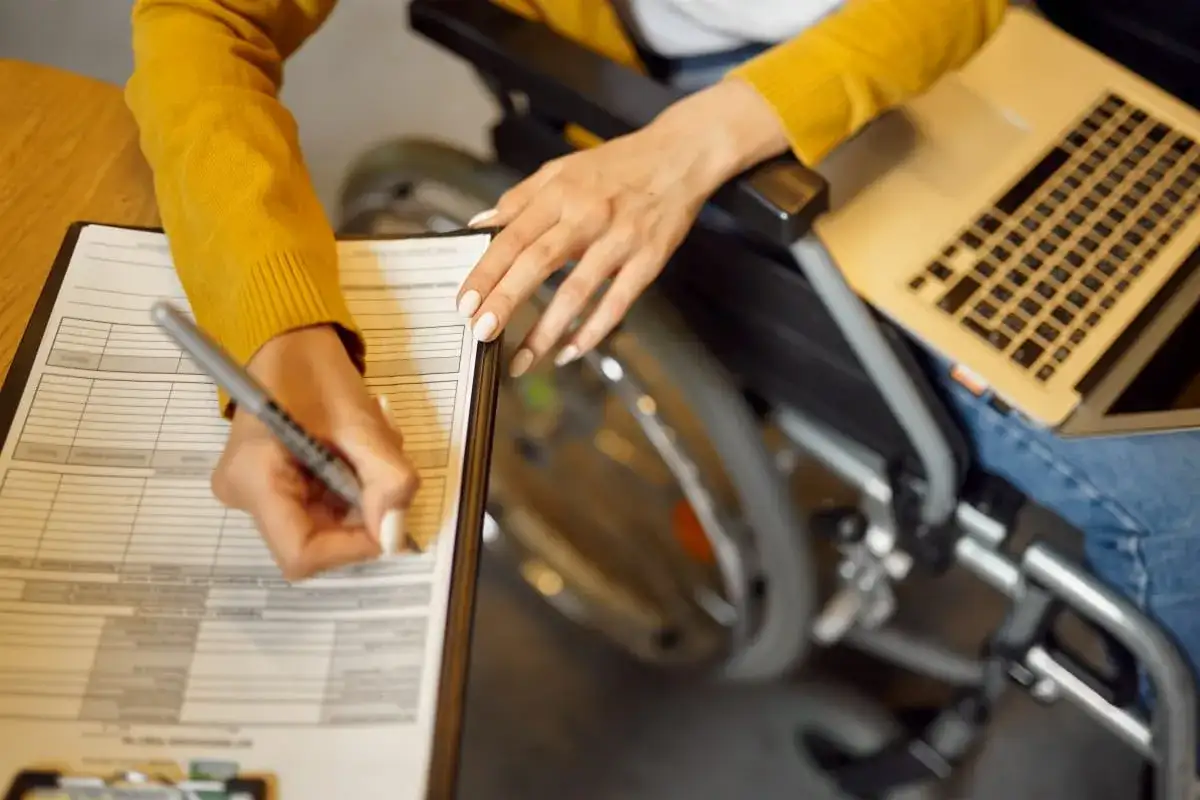 Kobieta na wózku inwalidzkim wypełnia dokumenty, korzystając z laptopa. To może być wniosek o 500 plus dla niepełnosprawnych.