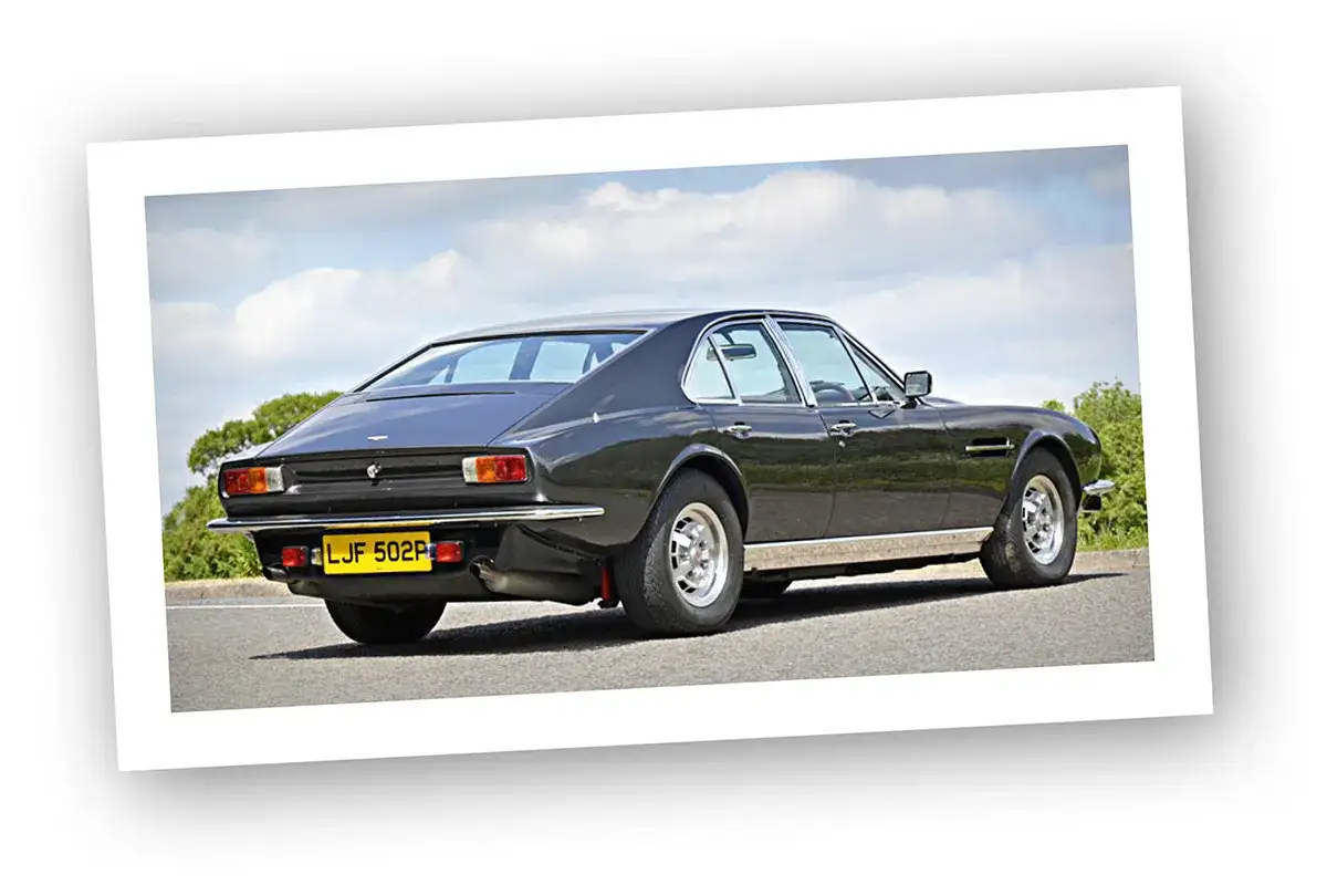 Klasyczny Aston Martin Lagonda w ciemnym kolorze, widziany z tyłu, na tle błękitnego nieba z chmurami.