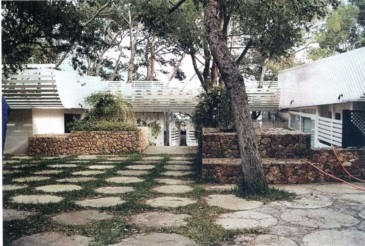 La casa Huarte, rodeada de árboles, presenta un patio con círculos de piedra y césped, muros de piedra y un diseño arquitectónico moderno.