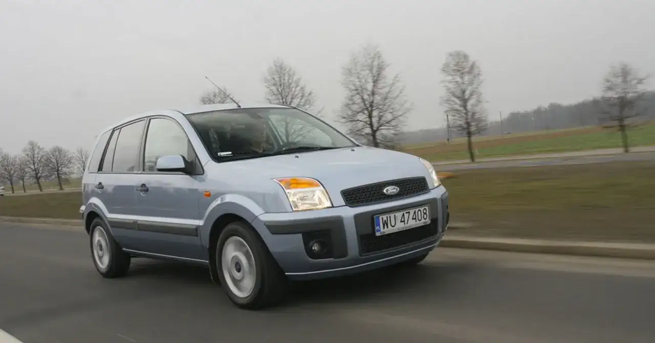 Jasnoszary Ford Fusion 1.4 benzyna jedzie po drodze. W tle drzewa i pola.