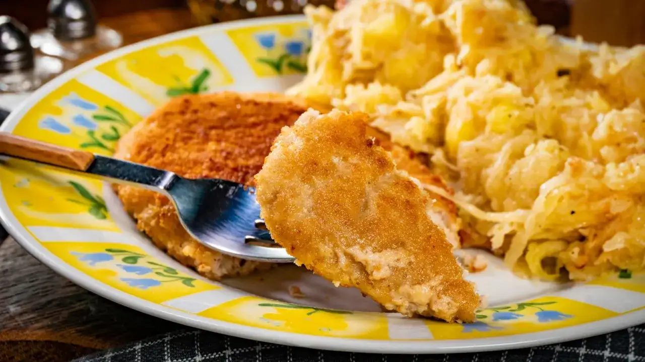Złocisty kotlet po parysku, chrupiący z wierzchu, z widelcem wbitym w kawałek. Obok porcja ziemniaków.