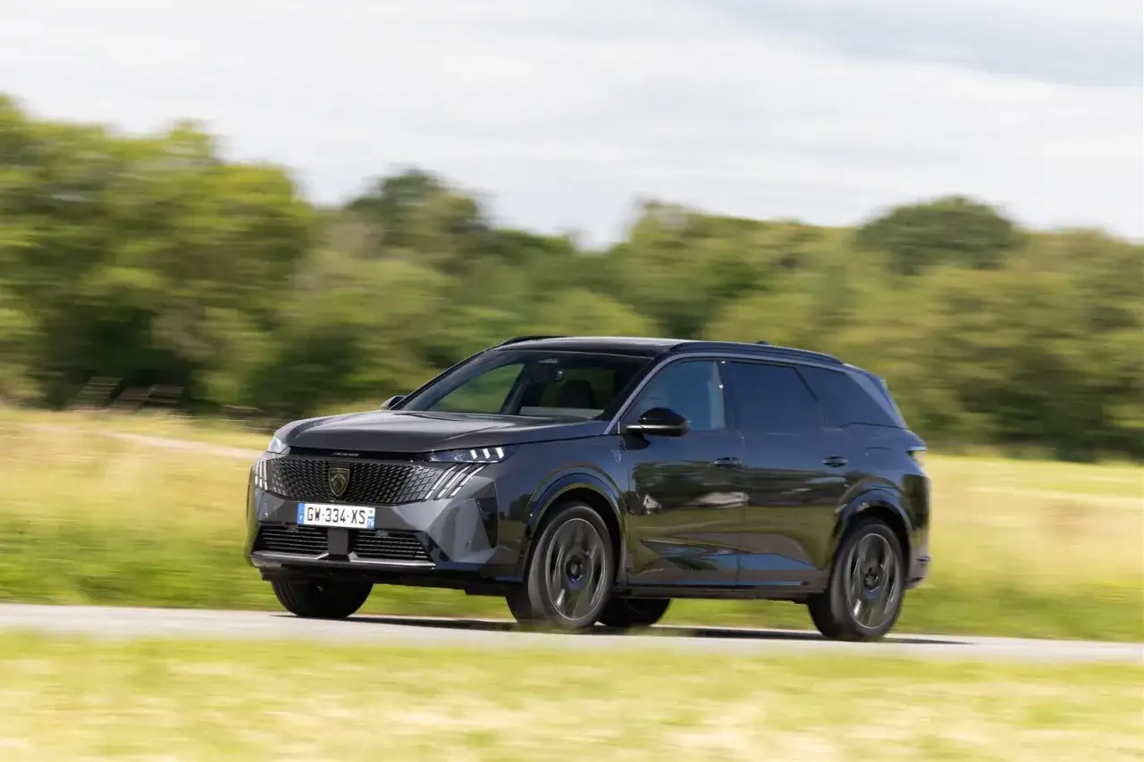 Ciemnoszary Peugeot 5008 jedzie po drodze wśród zielonych pól i drzew.
