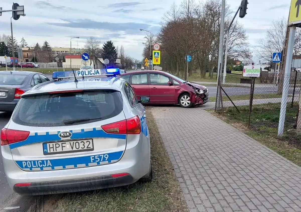 Samochód policyjny z włączonymi sygnałami świetlnymi przy drodze, gdzie doszło do kolizji drogowej. Czerwony samochód ma uszkodzony przód.