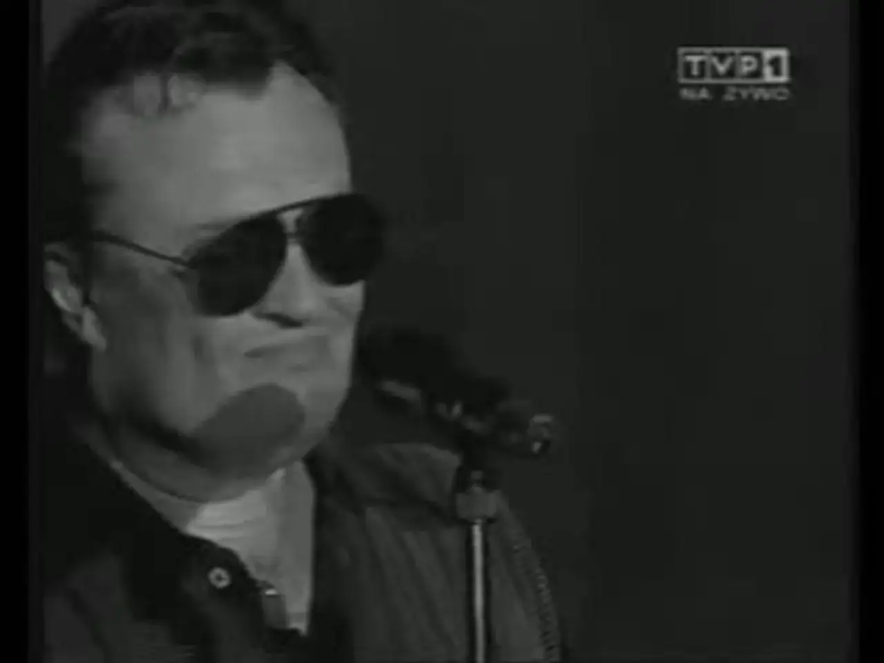 Mężczyzna w okularach przeciwsłonecznych śpiewa do mikrofonu. Widać logo TVP1. A po nocy przychodzi dzień.