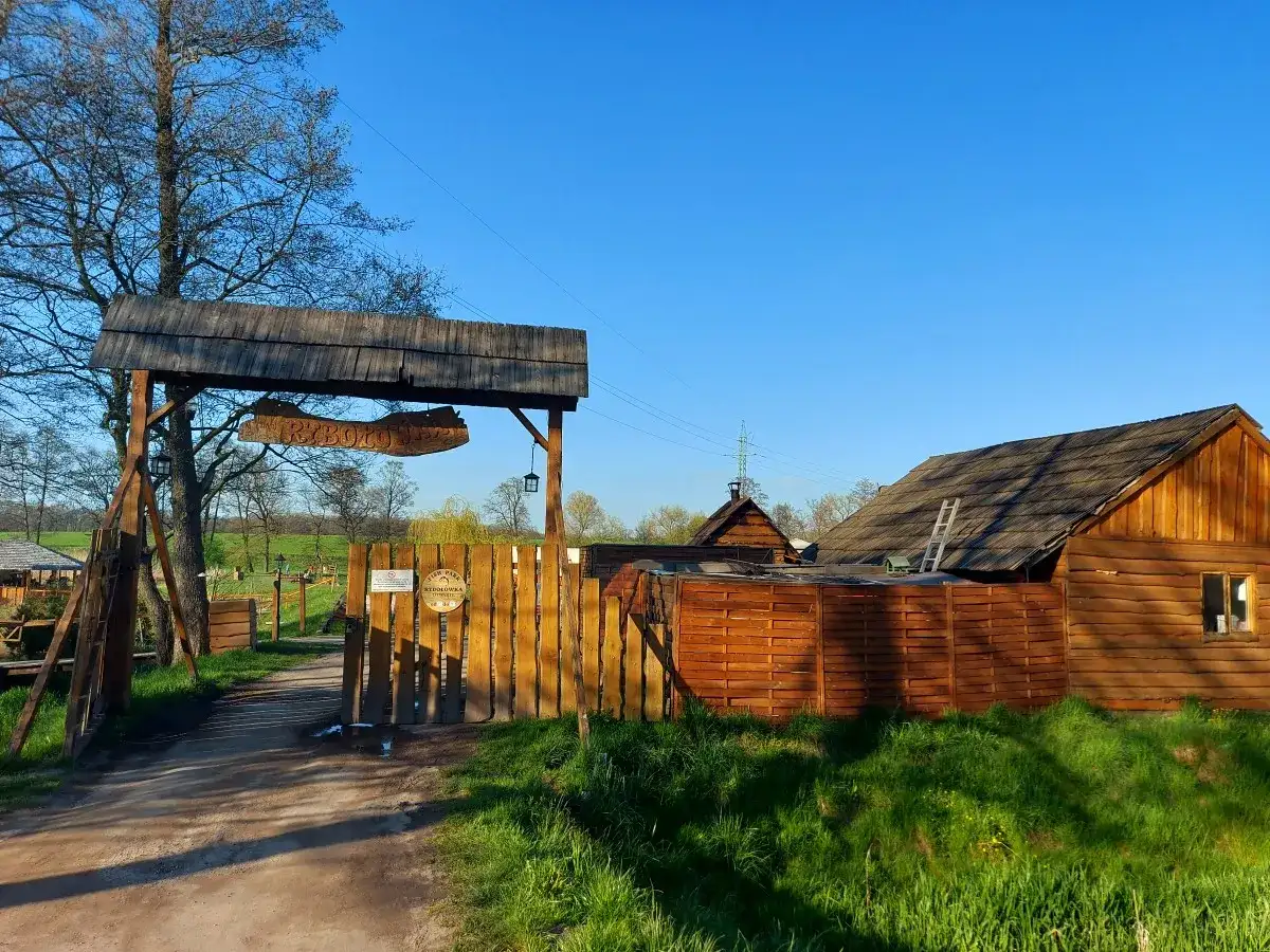 Drewniana brama z napisem "RYBOŁÓWKA" prowadzi do obiektu w Mikołowie. W tle widać chaty i zielone łąki.