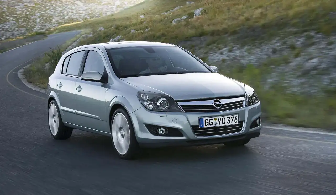 Srebrny Opel Astra 2005 na krętej drodze, otoczony zielonymi wzgórzami.