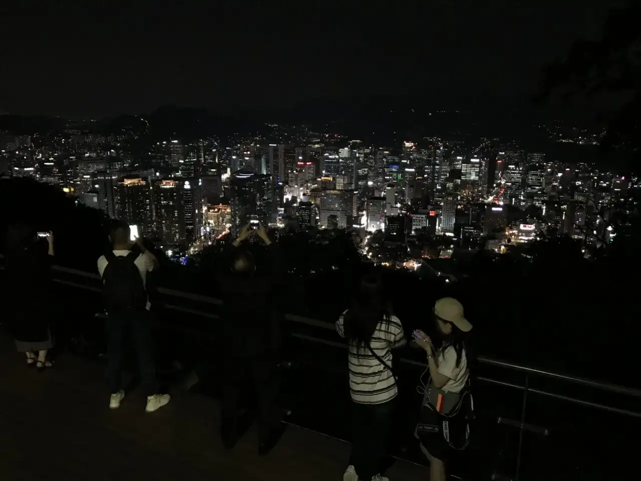 Nocny widok na rozświetlone miasto, przypominające panoramę Gangnam. Ludzie robią zdjęcia, podziwiając miejski krajobraz.