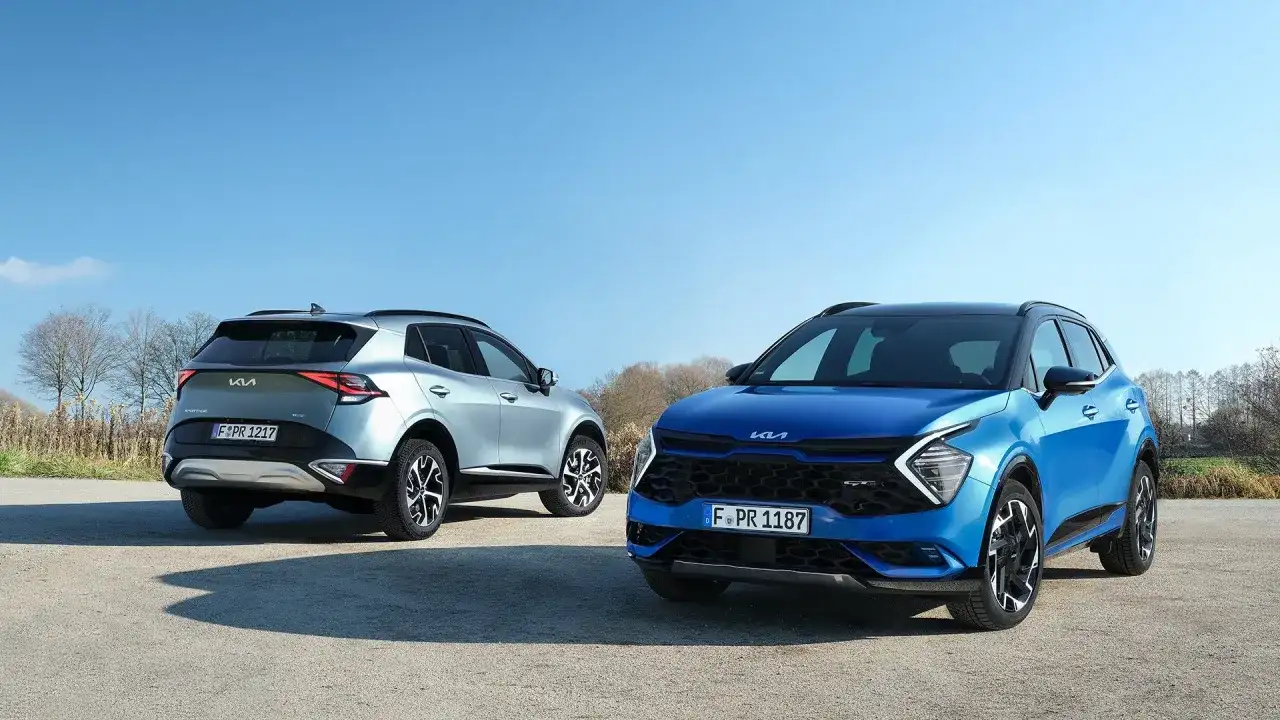 Dwa Kia Sportage GT Line: jeden niebieski z przodu, drugi srebrny z tyłu, na tle błękitnego nieba i drzew.