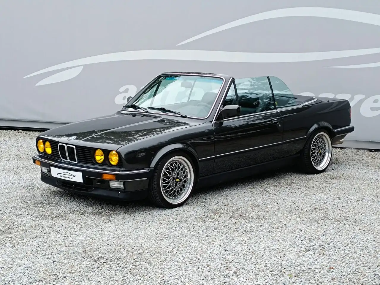 Czarny kabriolet BMW E30 z żółtymi światłami i felgami BBS. Klasyk na drodze.