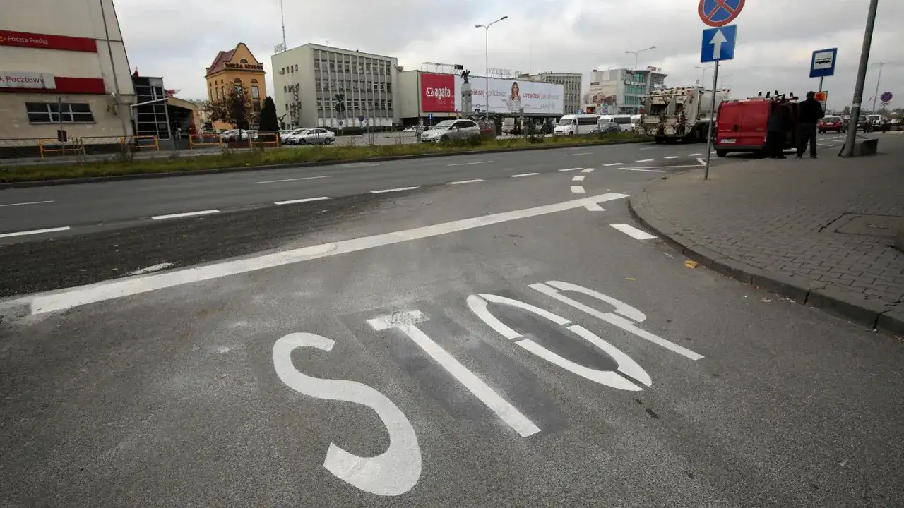 Na asfalcie widoczna jest biała linia warunkowego zatrzymania "STOP". W tle budynki i ruch uliczny.