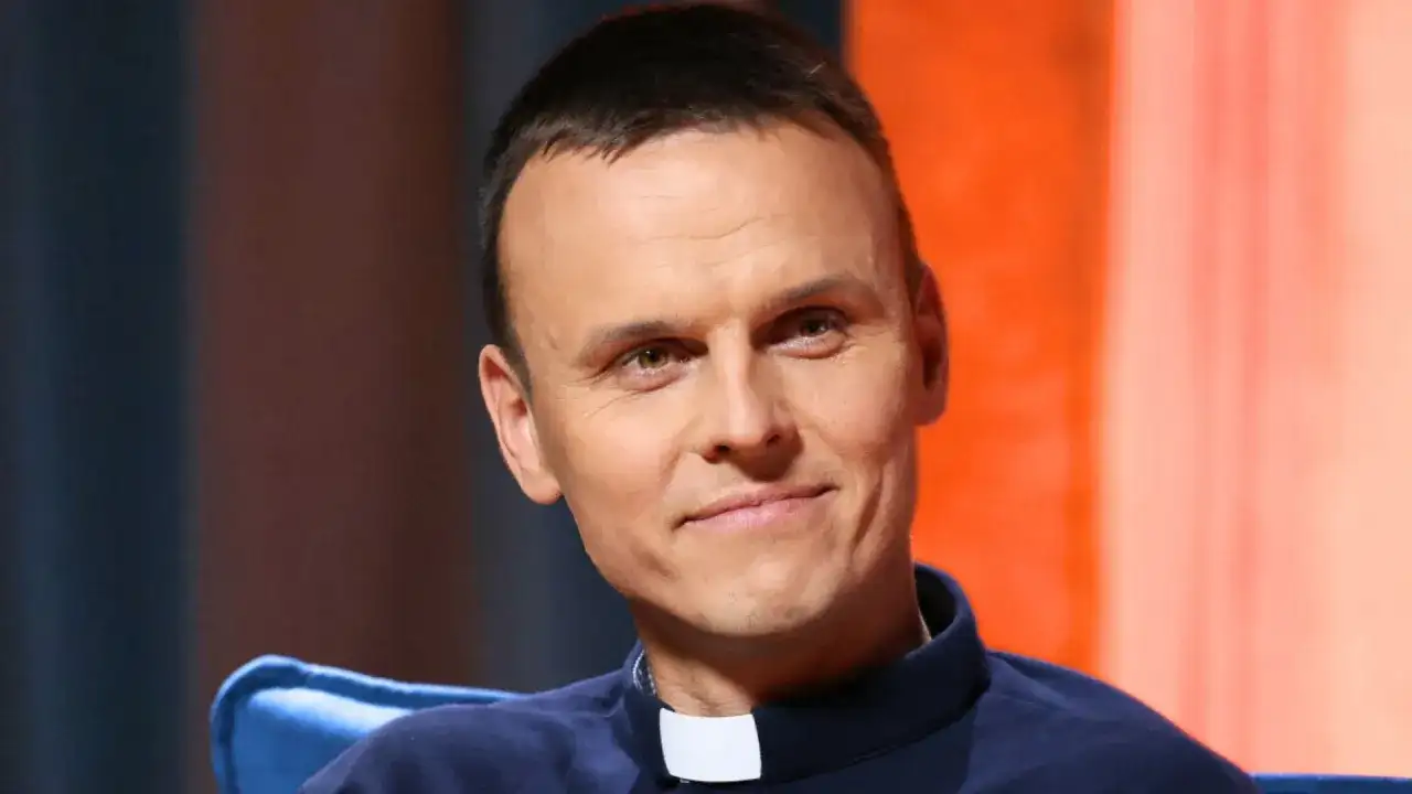 Uśmiechnięty ksiądz z osiedla, młody duchowny w sutannie, z uwagą słucha rozmówcy.