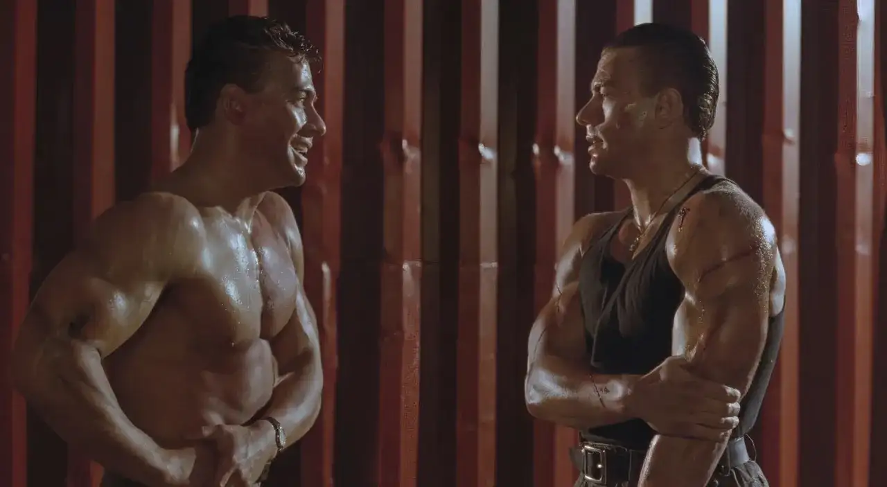 Dwaj Jean-Claude Van Damme w scenie z jednego z jego filmów, spoceni i z widocznymi ranami, patrzą na siebie.