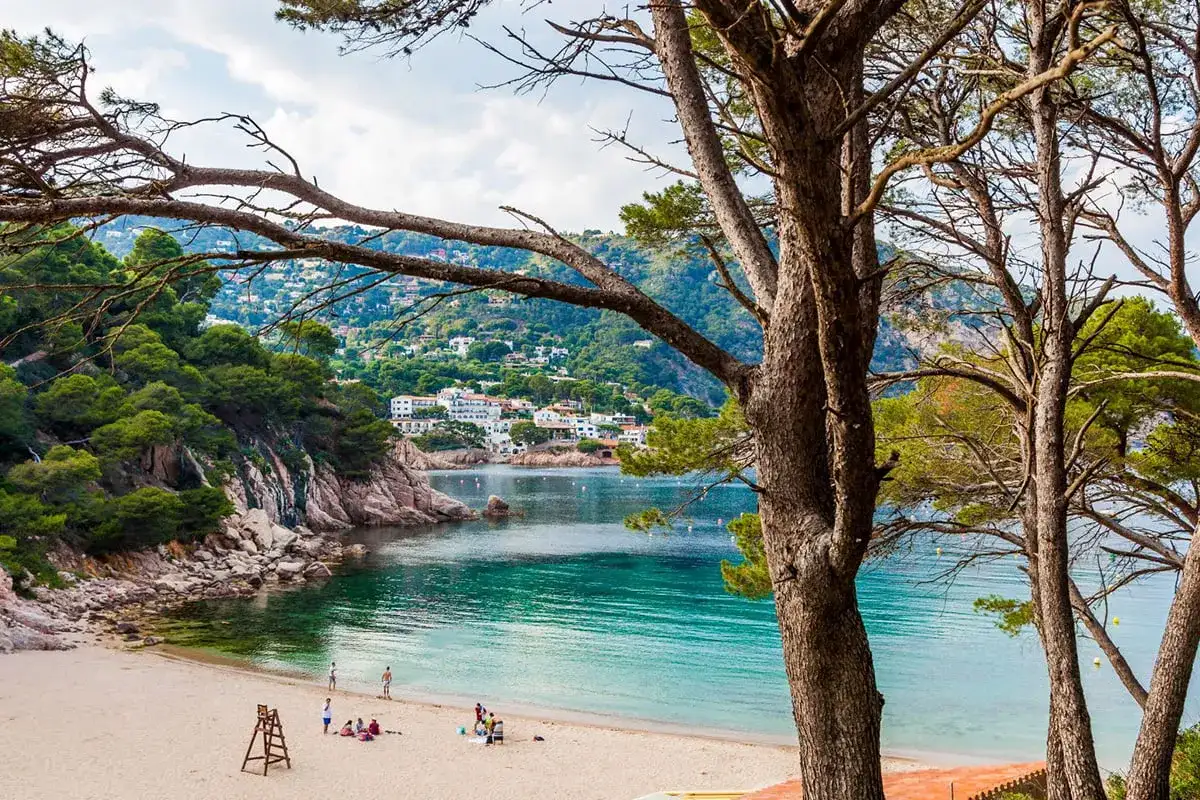 Piękna plaża w Hiszpanii z turkusową wodą, piaszczystym brzegiem i zielonymi wzgórzami w tle. Kilka osób odpoczywa na piasku.