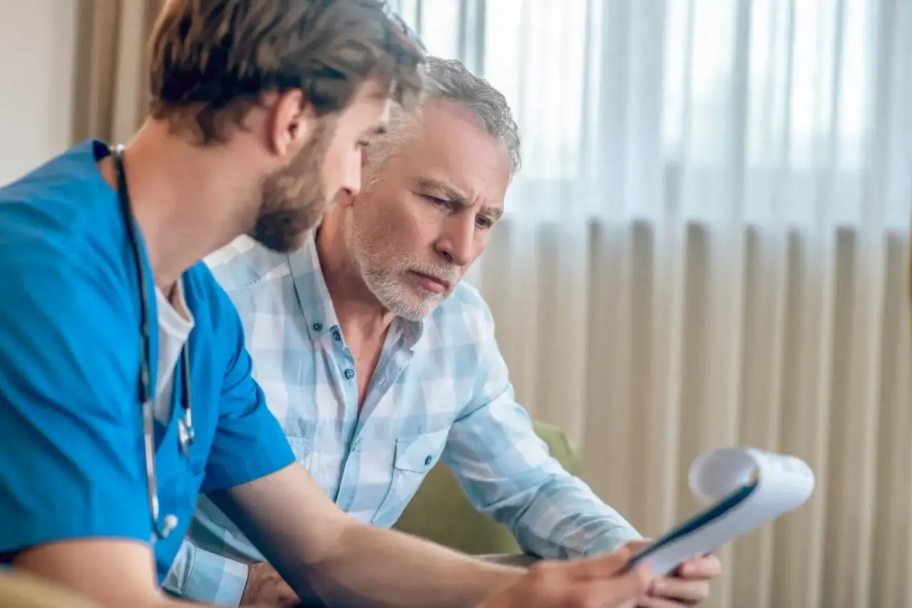 Lekarz w niebieskim fartuchu omawia wyniki badań z pacjentem. Mężczyzna z siwą brodą uważnie słucha, analizując dokumenty dotyczące biopsji prostaty.