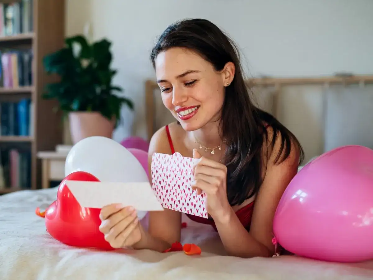 Uśmiechnięta kobieta czyta kartkę z serduszkami, otoczona balonami. Może to być inspiracja na wiersze dla ukochanego mężczyzny.