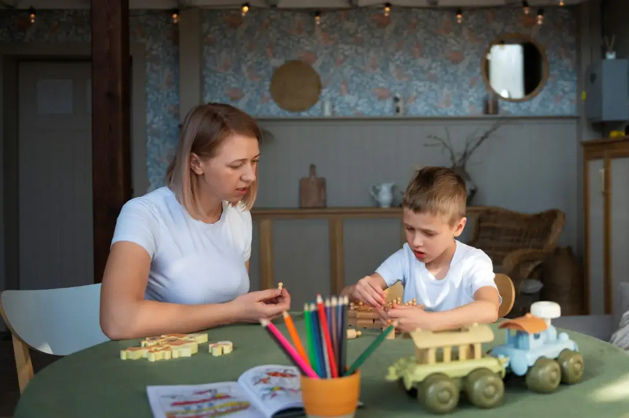 Kobieta i chłopiec grają w drewniane gry na zielonym stole. W tle widać wystrój pokoju, przypominający salę w przedszkolu niepublicznym.