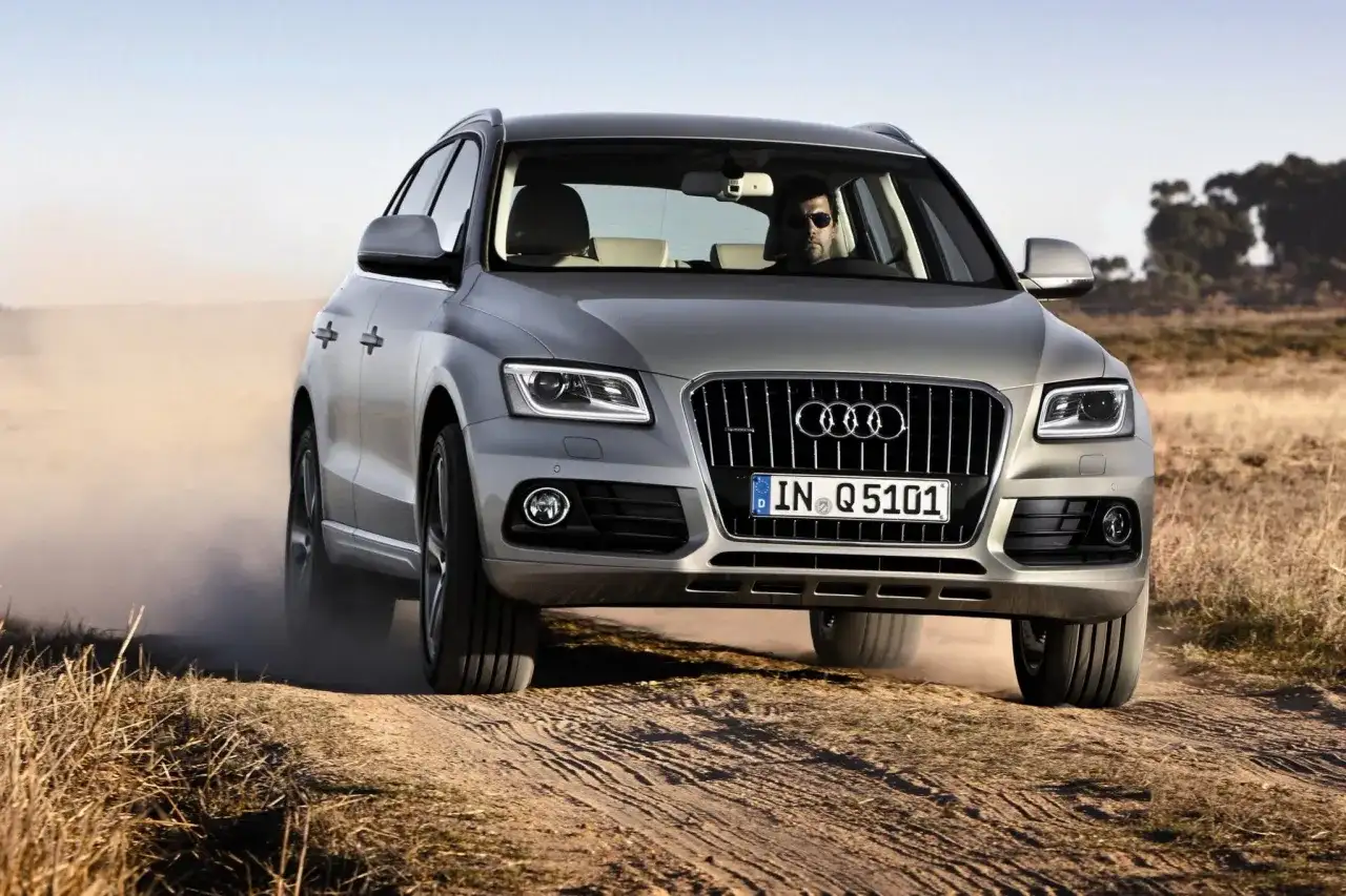 Srebrny Audi Q5 z kierowcą w okularach jedzie po piaszczystej drodze, wzbijając kurz. Wiele osób szuka audi q5 opinie przed zakupem.