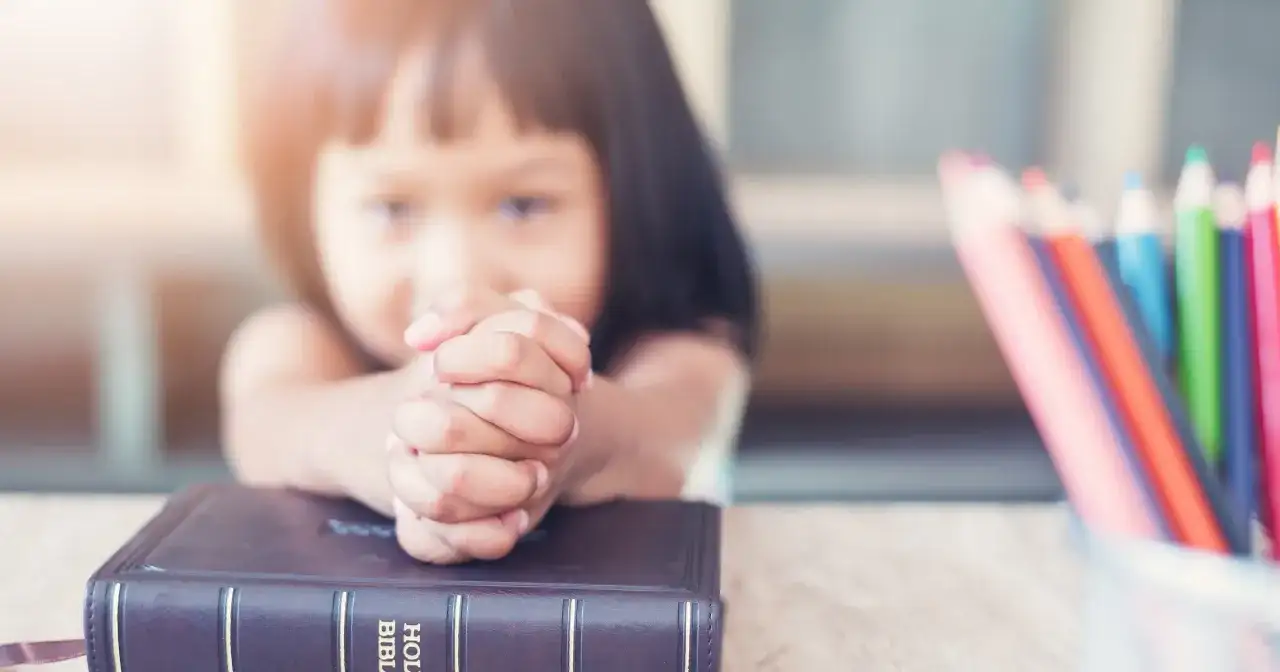 Dziecko z modlącymi się dłońmi na Biblii, obok kolorowe kredki. Nauka 10 przykazań bożych dla dzieci.