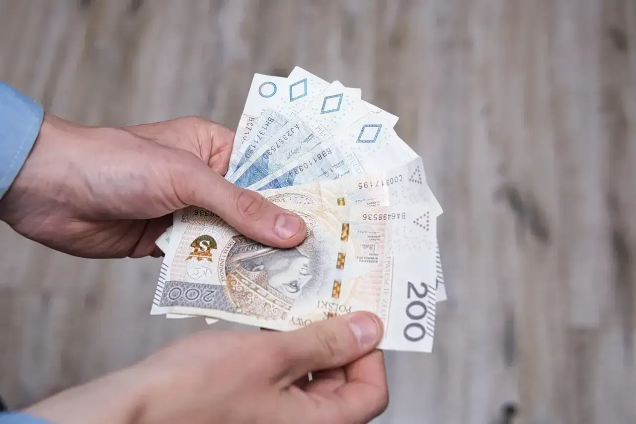 Dłonie trzymają polskie banknoty, w tym 200 zł. Może to symbolizować otrzymanie renty socjalnej z MOPS.