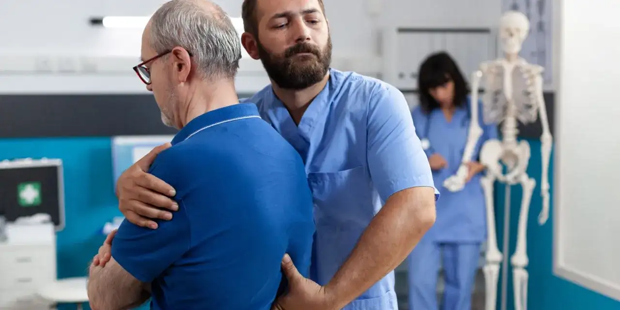 Fizjoterapeuta pomaga starszemu mężczyźnie z bólem pleców. W tle widać szkielet i drugą pielęgniarkę.