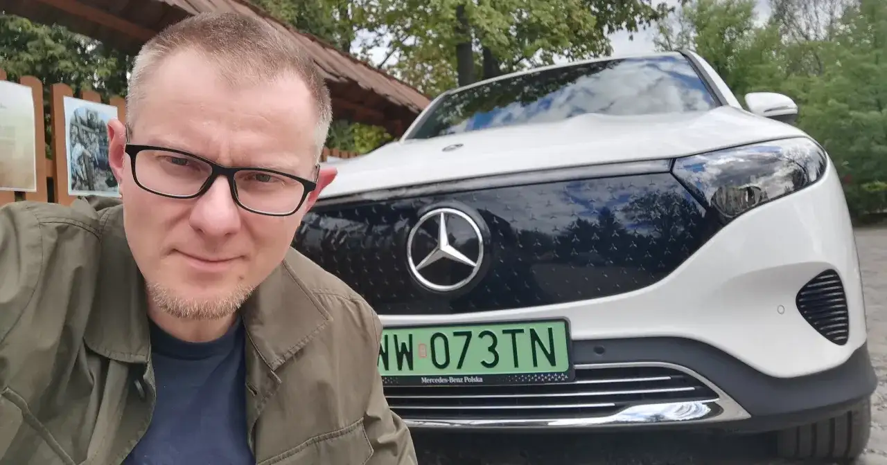 Mężczyzna w okularach pozuje do selfie na tle białego Mercedesa EQB.