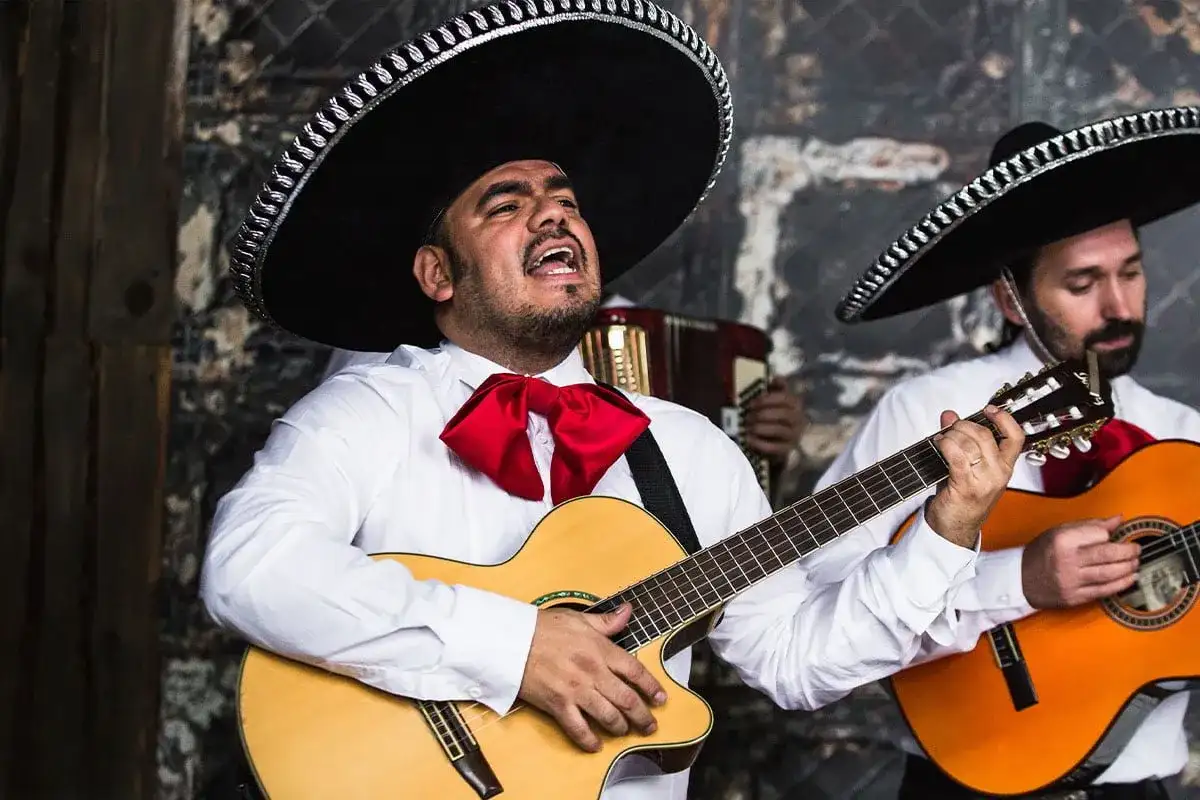 Zespół mariachi w tradycyjnych strojach, z wielkimi kapeluszami i czerwonymi muszkami, gra na gitarach i akordeonie.
