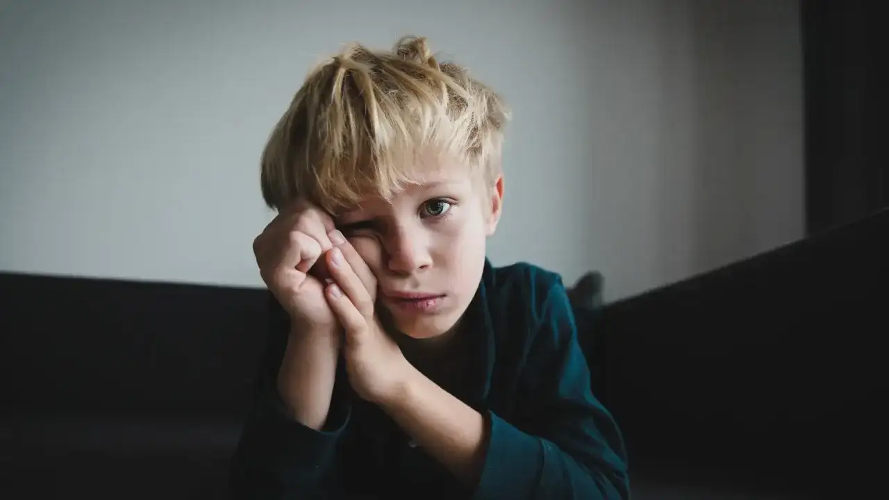 Chłopiec z blond włosami, zamyślony, opiera głowę na dłoniach. Jego spojrzenie sugeruje trudności, być może związane z zespołem Aspergera.