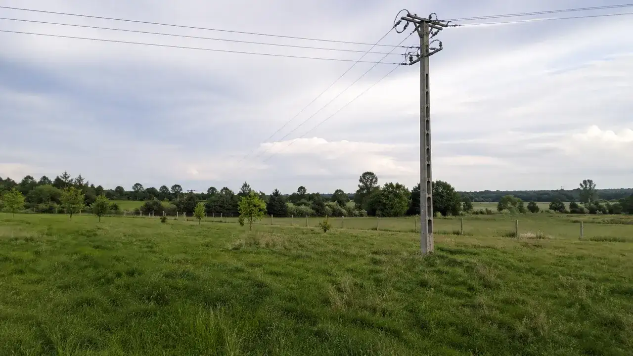 Słup na działce ponad 30 lat, stoi pośród zielonej łąki, z liniami energetycznymi biegnącymi ku horyzontowi.