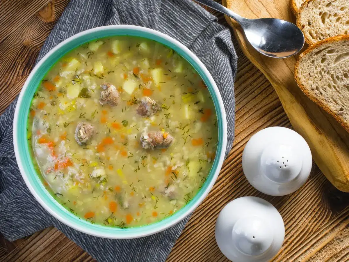 Aromaticzny krupnik, przepis babci, z kawałkami mięsa, ziemniaków i marchewki, posypany koperkiem. Obok kromki chleba i solniczki.