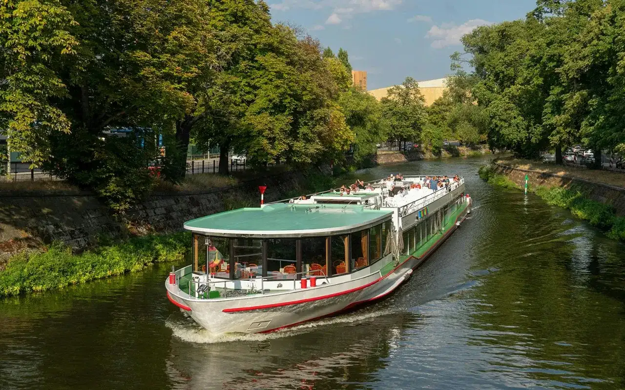 Statek wycieczkowy płynie po zielonej rzece w Berlinie, otoczony drzewami.