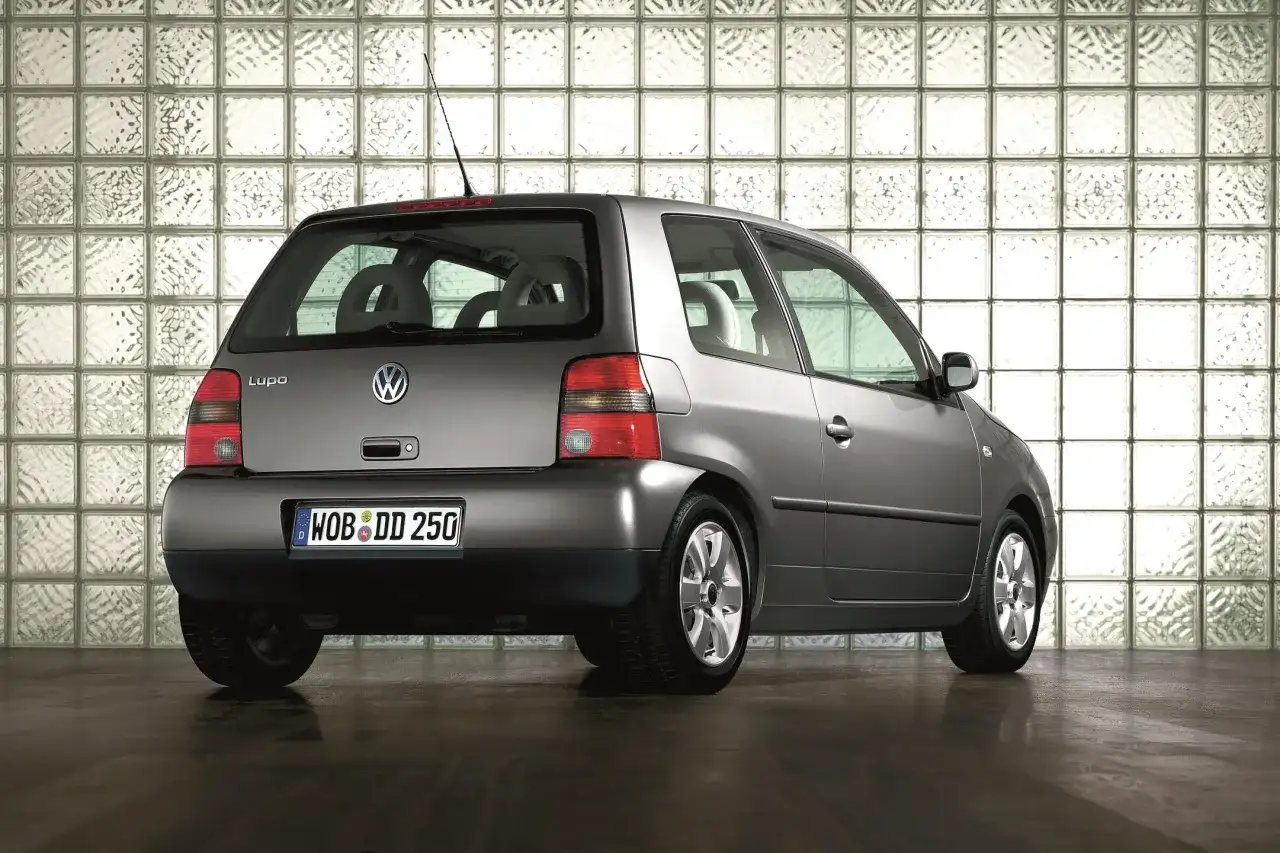 Szary Volkswagen Lupo stoi na ciemnej podłodze, na tle ściany z białych pustaków szklanych.
