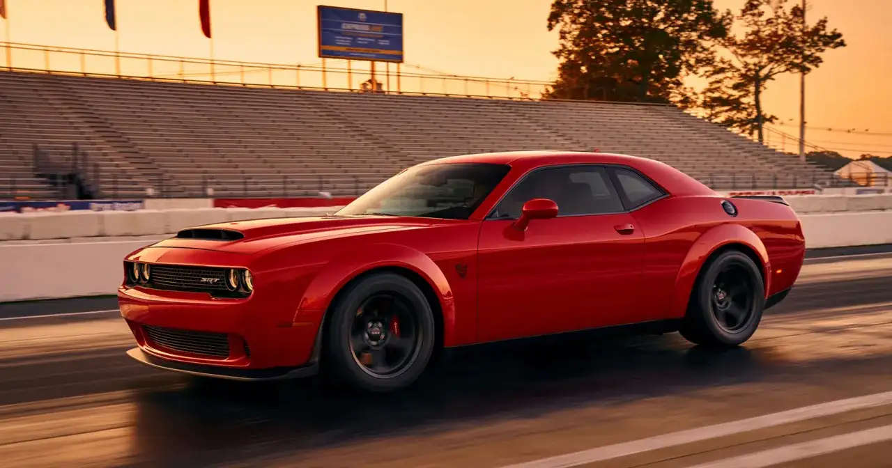Czerwony Dodge Demon pędzi po torze wyścigowym podczas zachodu słońca, z trybunami w tle.