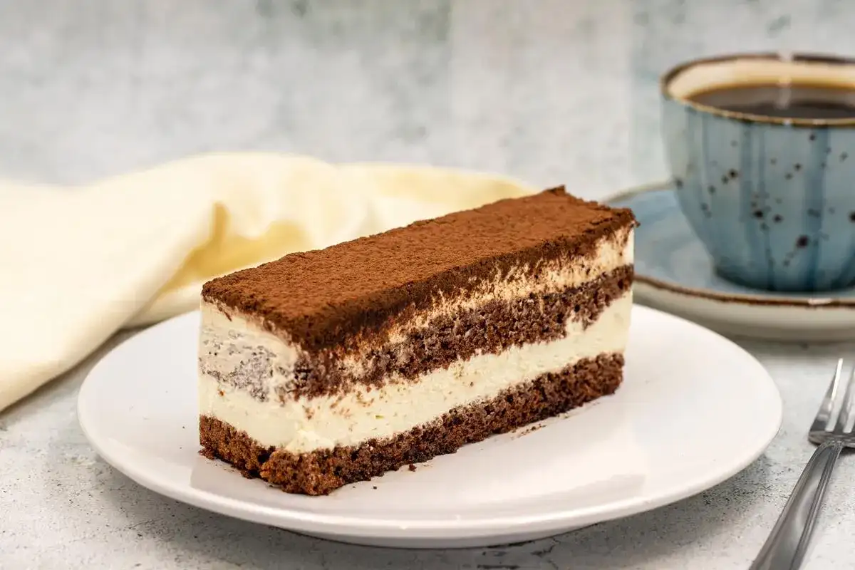Pyszne ciasto czekoladowe z mascarpone, posypane kakao, na białym talerzu, obok filiżanka kawy.