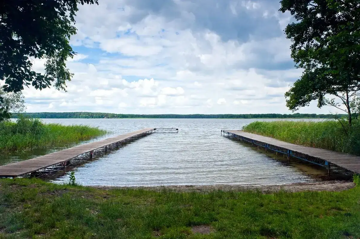 Dwa drewniane pomosty prowadzące na spokojne jezioro, otoczone zielenią. Idealne miejsce na relaks i podziwianie przyrody, jedna z wielu atrakcji w Boszkowie.