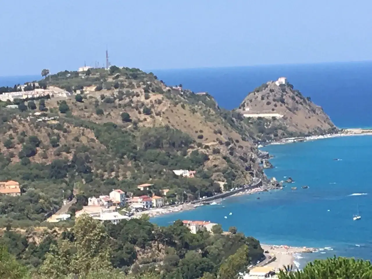 Panoramica di Barcellona Pozzo di Gotto con case bianche e rosse affacciate sul mare blu intenso, colline verdi e un castello in cima.