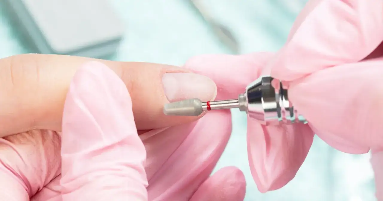Dłonie w różowych rękawiczkach wykonują manicure frezarką. Przygotowanie paznokci do porodu, by były schludne i bezpieczne.