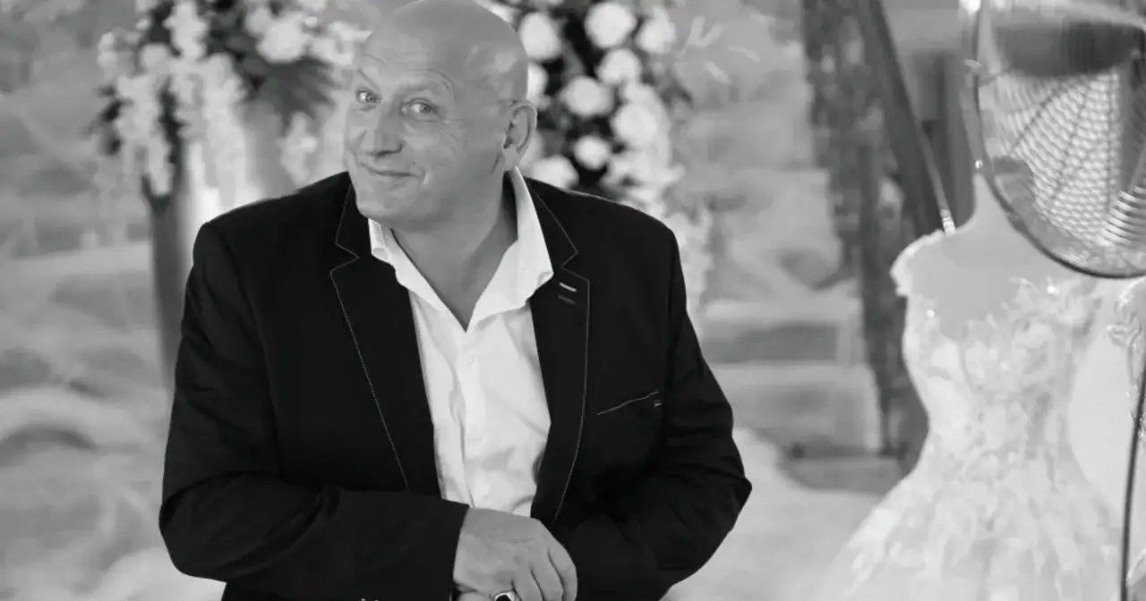Jacek, znany jako królowe życia, w eleganckim garniturze, z uśmiechem spogląda w obiektyw. W tle widać suknię ślubną.