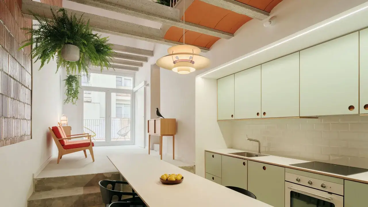 Interior de una casa entre medianeras con cocina moderna, sala de estar con sillón rojo y plantas colgantes.