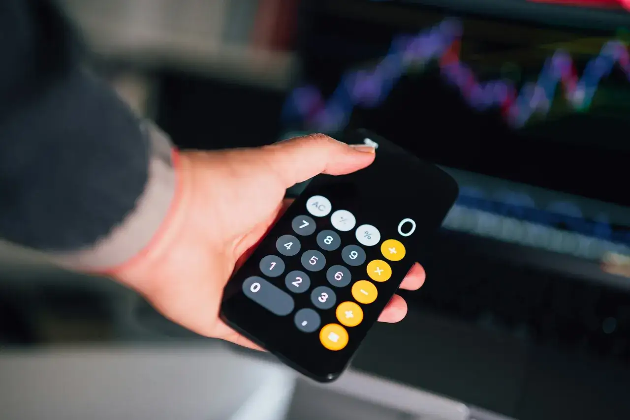 Dłoń trzyma smartfon z kalkulatorem, na ekranie widać wykresy finansowe. Obliczanie faktury bez vat.