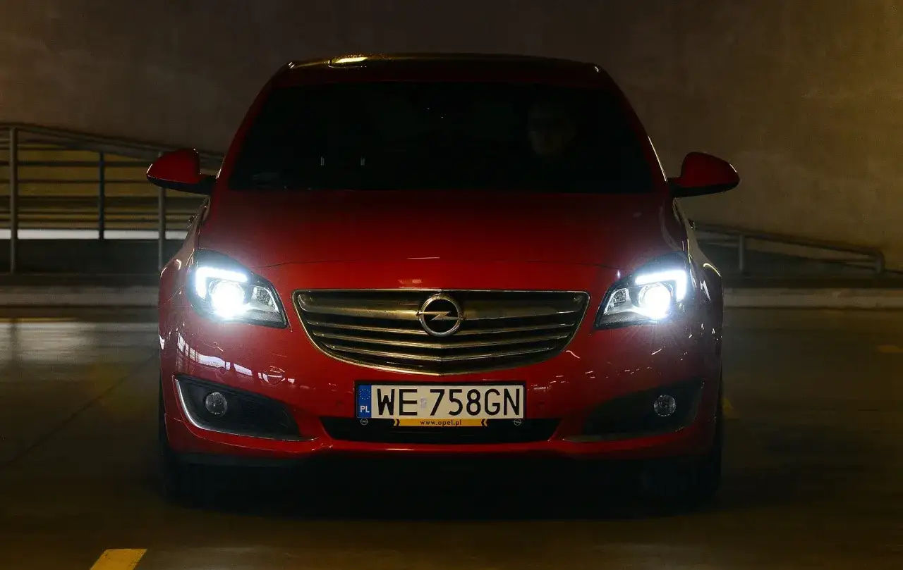 Czerwony Opel z włączonymi światłami, gotowy do jazdy. Dowiedz się, jak ustawić światła w samochodzie domowym sposobem, by zapewnić sobie bezpieczeństwo.