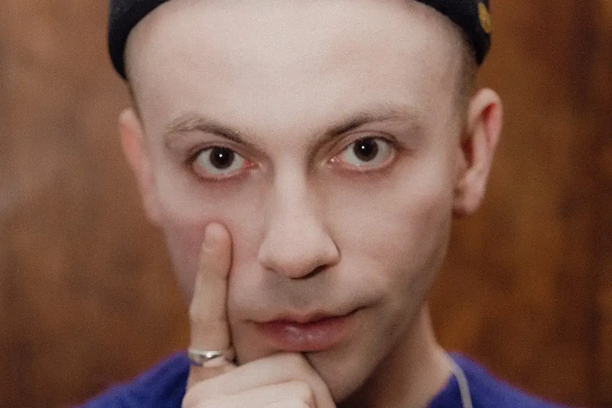Zbliżenie na twarz Arka Kłusowskiego w czarnej czapce, z palcem przy policzku.