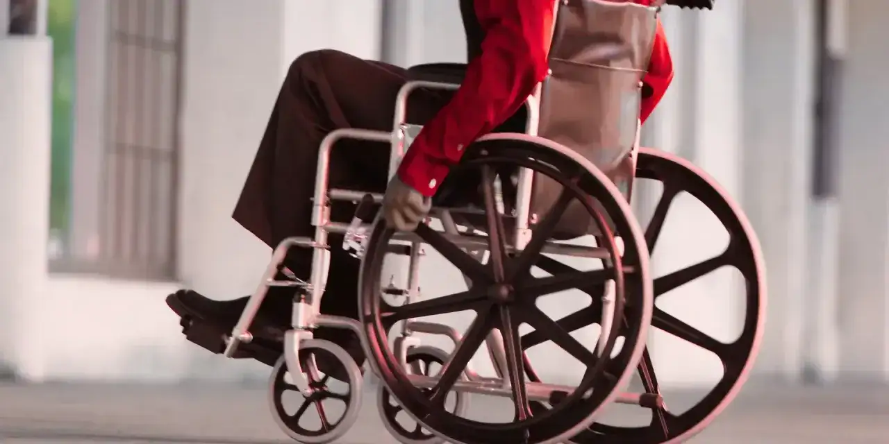 Osoba na wózku inwalidzkim w czerwonej koszuli i czarnych spodniach porusza się po chodniku.