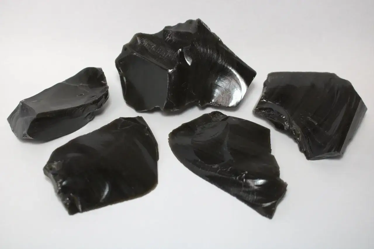 Fünf scharfkantige Stücke glänzenden, schwarzen Obsidiansteins liegen auf weißem Untergrund.